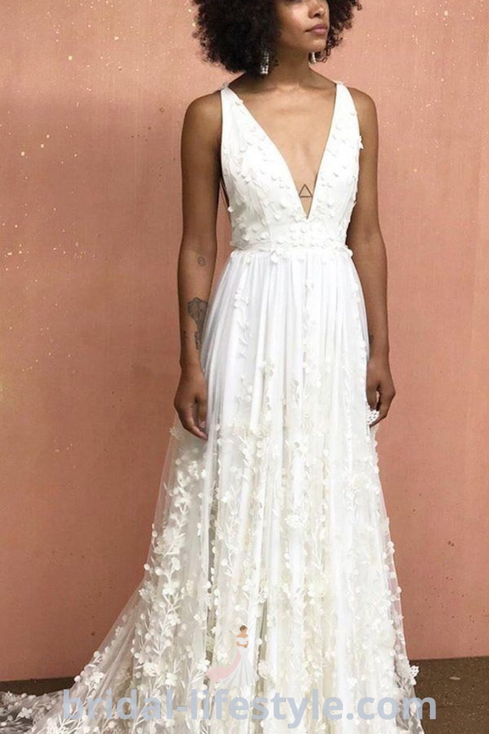 A petite woman in a white floral boho bridal dress stands gracefully in front of a pink wall, showcasing the perfect look for a dreamy beach wedding. This elegant gown represents the best trends for brides at bridal-lifestyle.com.