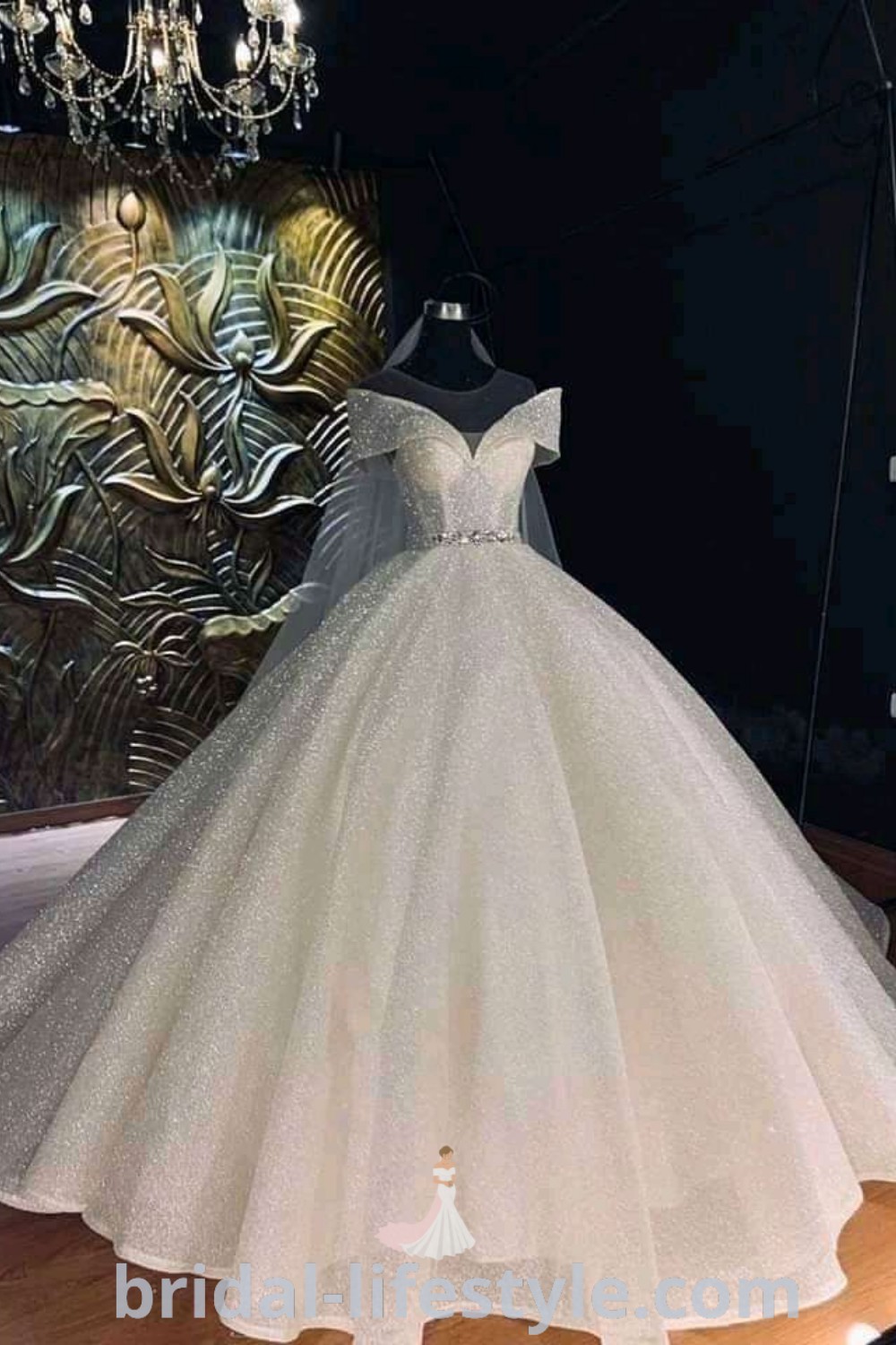 An elegant white wedding dress on display in front of a chandelier, showcasing a glitter ball gown style, perfect for brides seeking vintage charm and romantic bridal aesthetic ideas. Explore dresses of your dream at bridal-lifestyle.com.