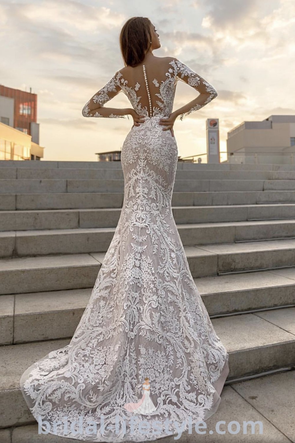 An elegant woman in a backless wedding dress with sleeves standing on steps, showcasing a romantic and stylish look for dreamy brides. This dress is a top choice for vintage wedding trends. Discover more bridal inspiration and dresses of your dreams at bridal-lifestyle.com.