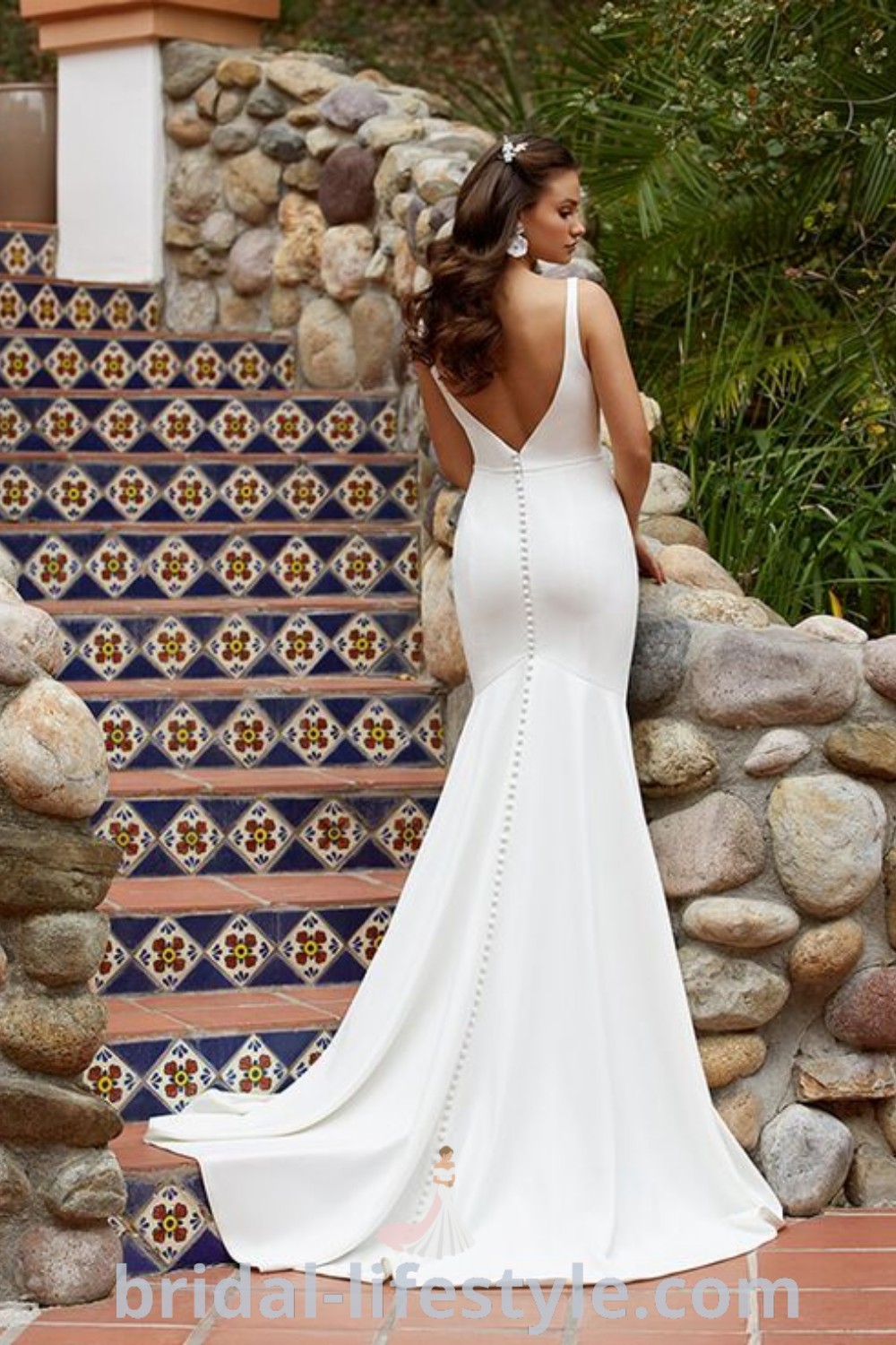 A woman in a white wedding dress with beaded details stands in front of a stone wall, embodying elegant bridal trends for a romantic atmosphere. This dress offers inspiration for dreamy brides at bridal-lifestyle.com, showcasing dresses of your dreams and the best aesthetic ideas.