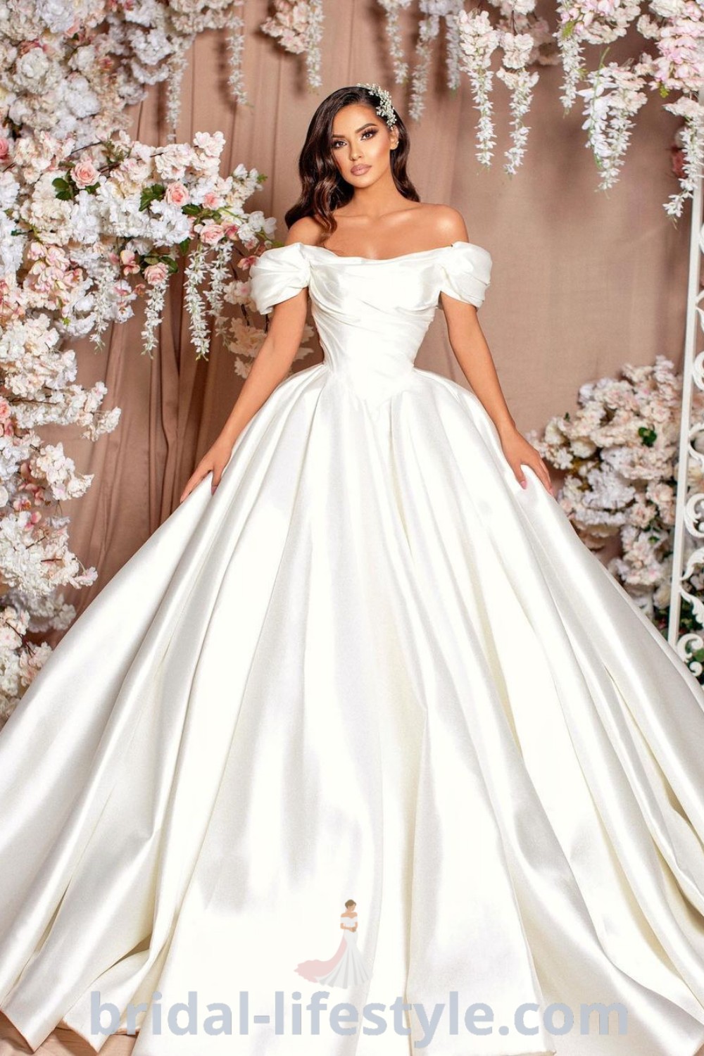 A woman in a white off the shoulder gown stands in front of a floral backdrop, showcasing a blend of elegance and romantic style. This dress represents the best trends in wedding attire for dreamy brides, perfect for their special day. Discover more at bridal-lifestyle.com.