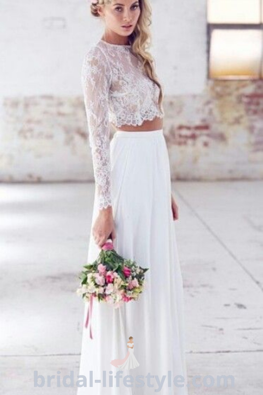 A woman wearing a vintage two-piece wedding dress with lace top, holding a bouquet, standing next to a brick wall. This elegant attire reflects bohemian wedding dress trends and offers aesthetic ideas for dreamy brides. Find more inspiration at bridal-lifestyle.com.