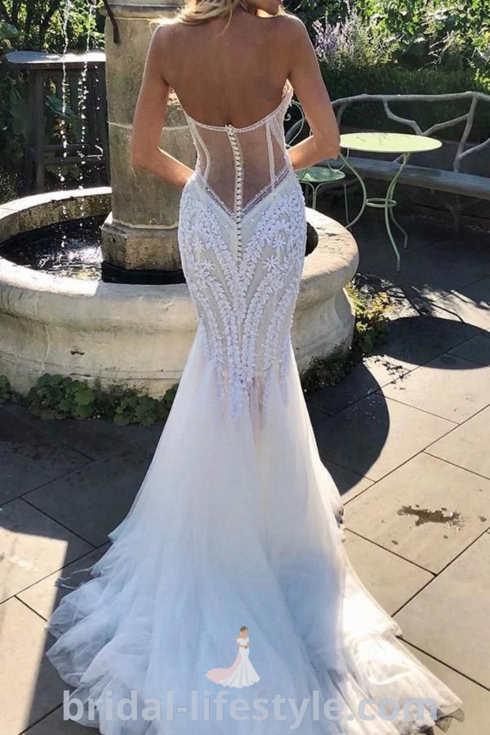 An elegant woman in a Pallas Couture wedding dress stands near a fountain, showcasing a unique wedding gown ideal for romantic and rustic weddings. This white wedding dress represents the best bridal trends and aesthetic ideas. Discover more inspiration for your special day at bridal-lifestyle.com.