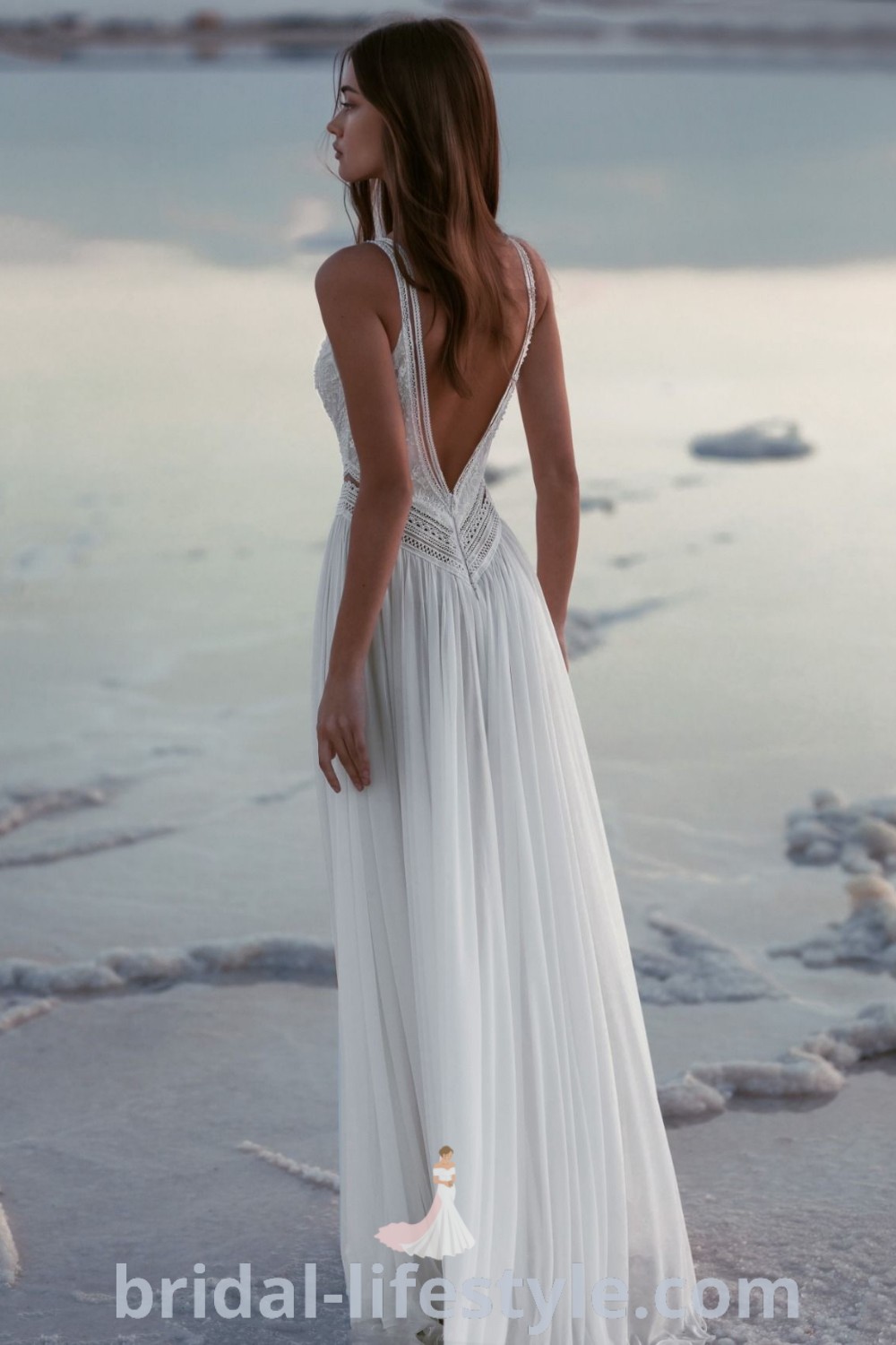 An elegant woman in a long white dress stands on the beach, showcasing a stunning beach wedding dress ideal for dreamy brides. This boho style aligns with current trends and offers inspiration for short beach wedding dresses and lace details. Perfect for destination wedding photography, find your ideal bridal look at bridal-lifestyle.com.