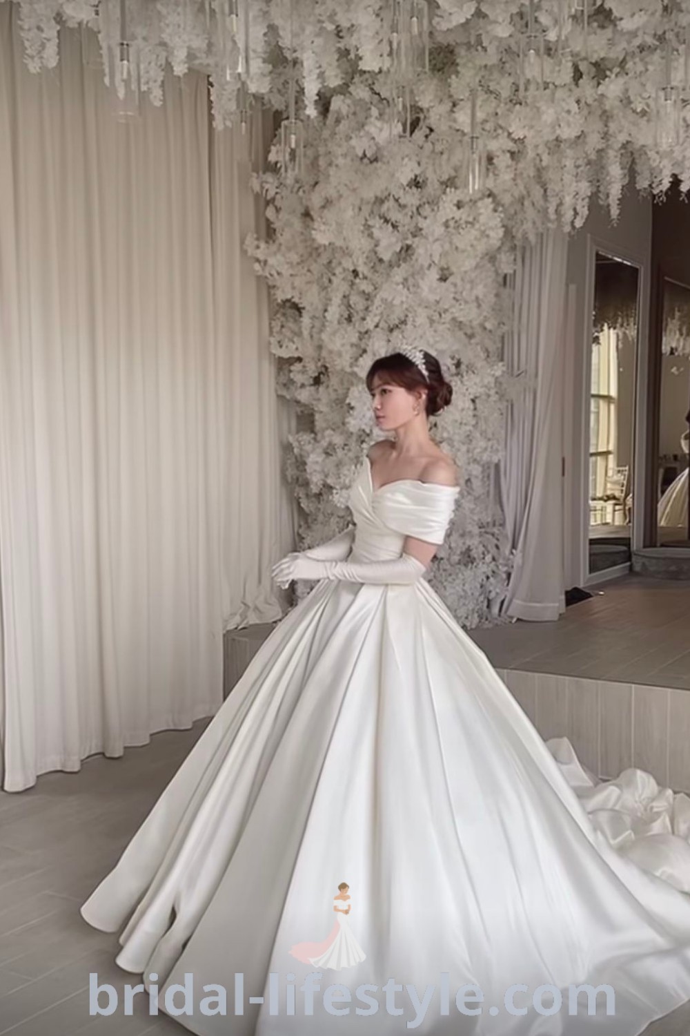 An elegant satin wedding dress with gloves and a train, paired with a flower-covered wall, offers inspiration for dreamy brides seeking fancy wedding dresses. Discover the best bridal aesthetic ideas at bridal-lifestyle.com.