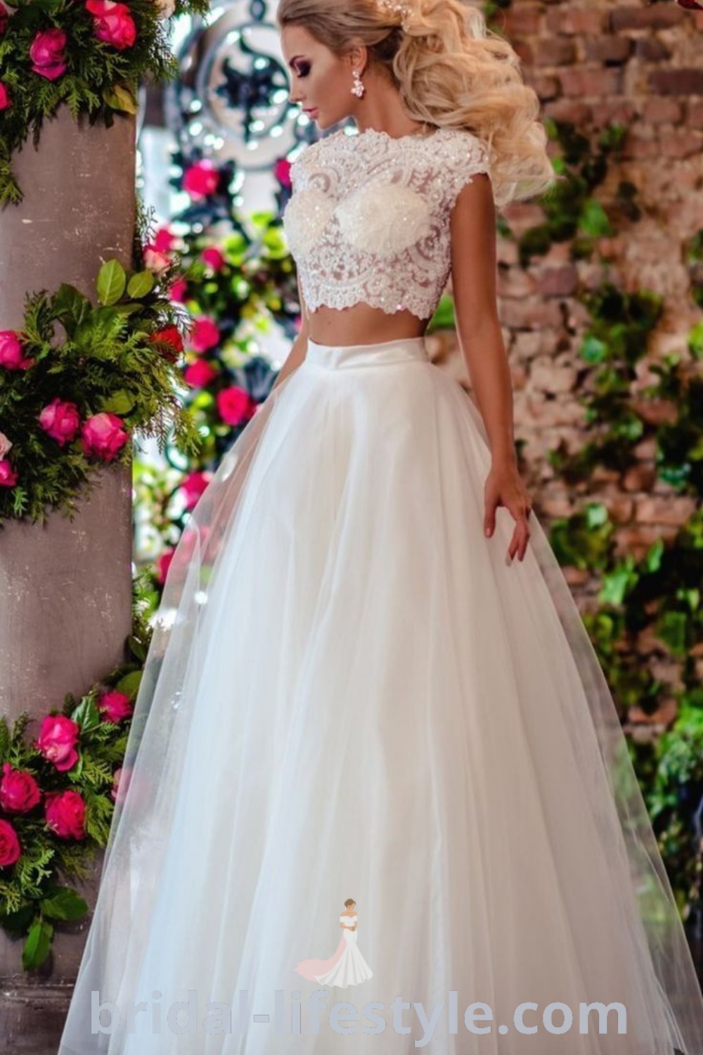 A woman in a white wedding dress standing next to a flower covered archway with pink roses, showcasing a cropped wedding dress two pieces. This elegant two piece wedding dress is perfect for dreamy brides, offering romantic inspiration and the best aesthetic ideas for the special day. Find more inspiration at bridal-lifestyle.com.