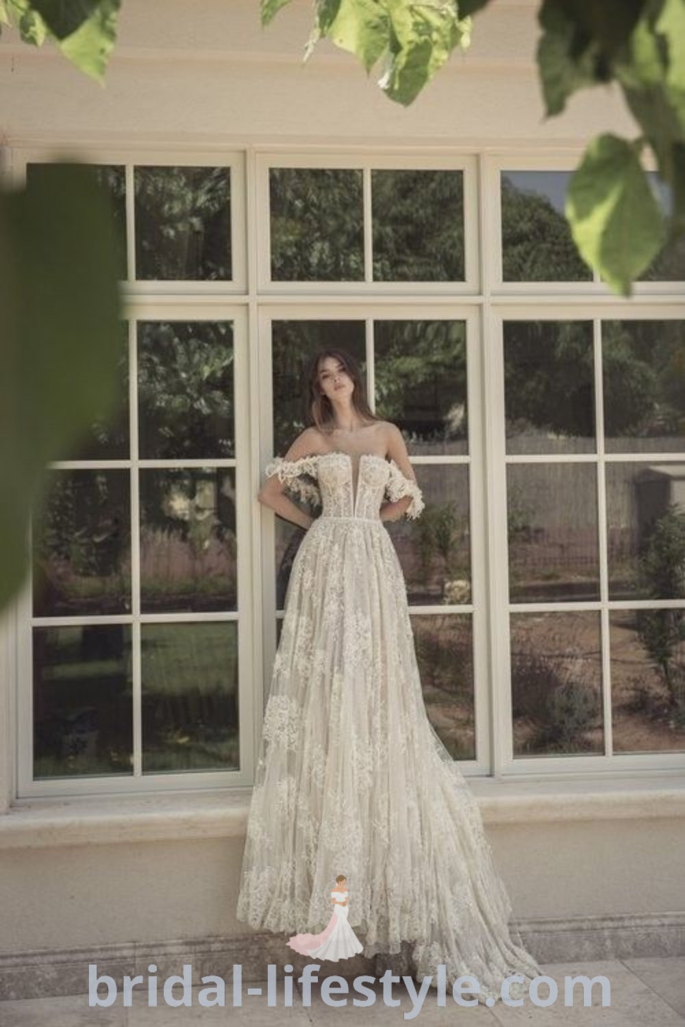 An elegant woman standing in front of a window wearing a wedding dress with an off-the-shoulder design, showcasing earthy and dark academia wedding styles. This long wedding dress features train fabric and reflects the latest bridal trends for dreamy brides. Find more inspiration ideas at bridal-lifestyle.com.