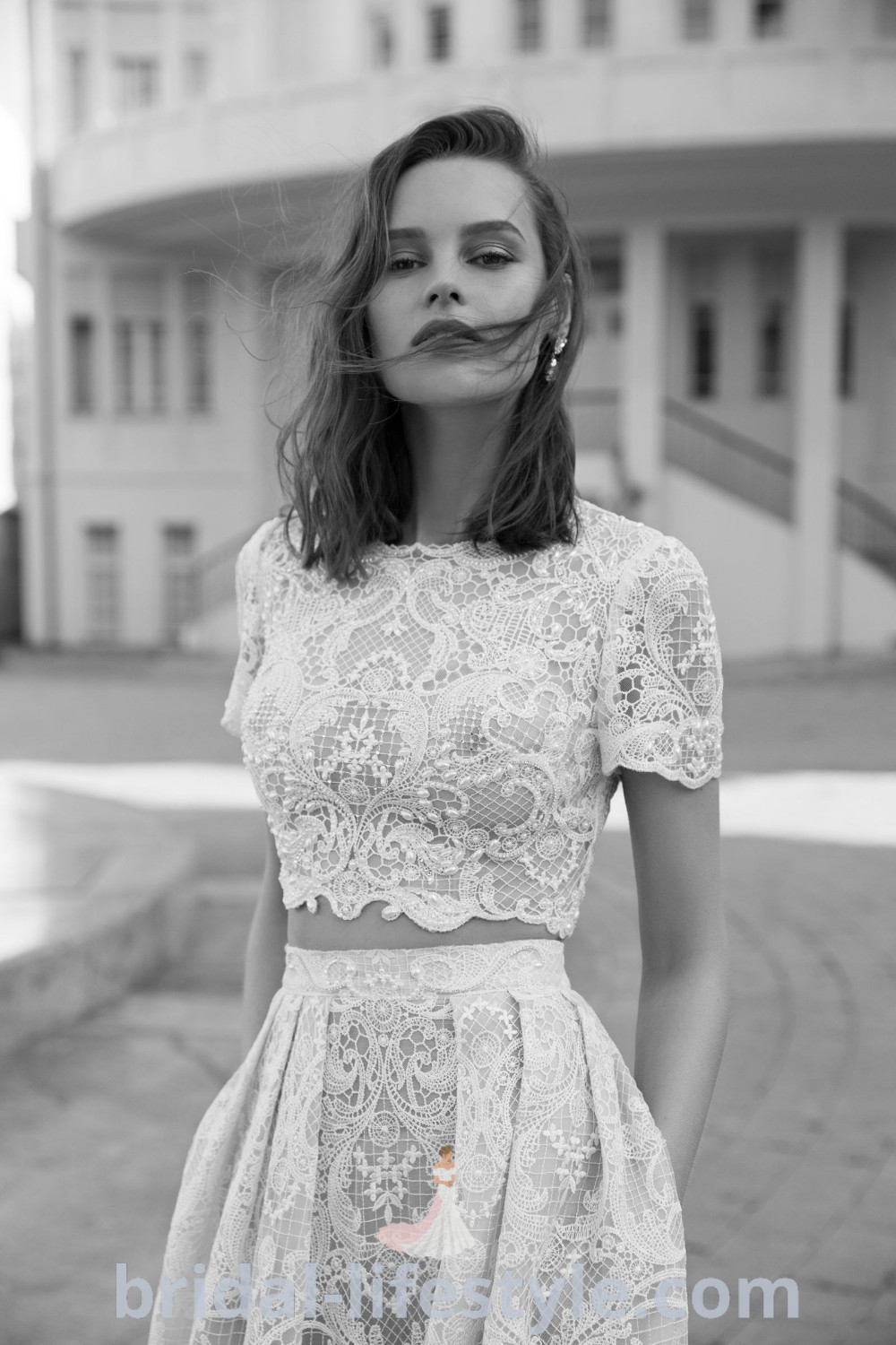 A woman standing in front of a building wearing an elegant bridal crop top and pants with a lace top, showcasing a stylish outdoor wedding outfit. This combination of the lace top and skirt embodies aesthetic ideas for every bride