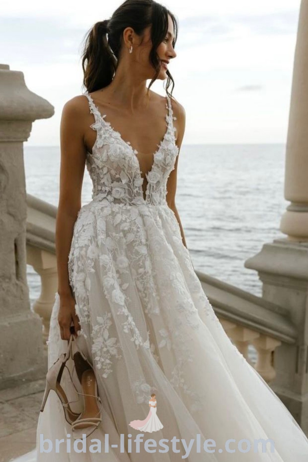 A woman in a white wedding dress with detachable sleeves stands on a balcony near the ocean, embodying coastal wedding dress inspiration. This elegant gown is perfect for tall brides and reflects the best aesthetic ideas for every bride’s unique taste. Discover more at bridal-lifestyle.com.