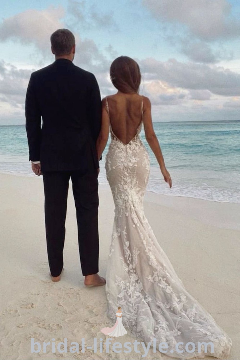 A man and woman stand on the beach in wedding dresses, looking out at the ocean, showcasing elegant beach wedding styles, including a backless mermaid dress. Perfect for dreamy brides, these dresses represent the best aesthetic ideas for a romantic wedding day at bridal-lifestyle.com.