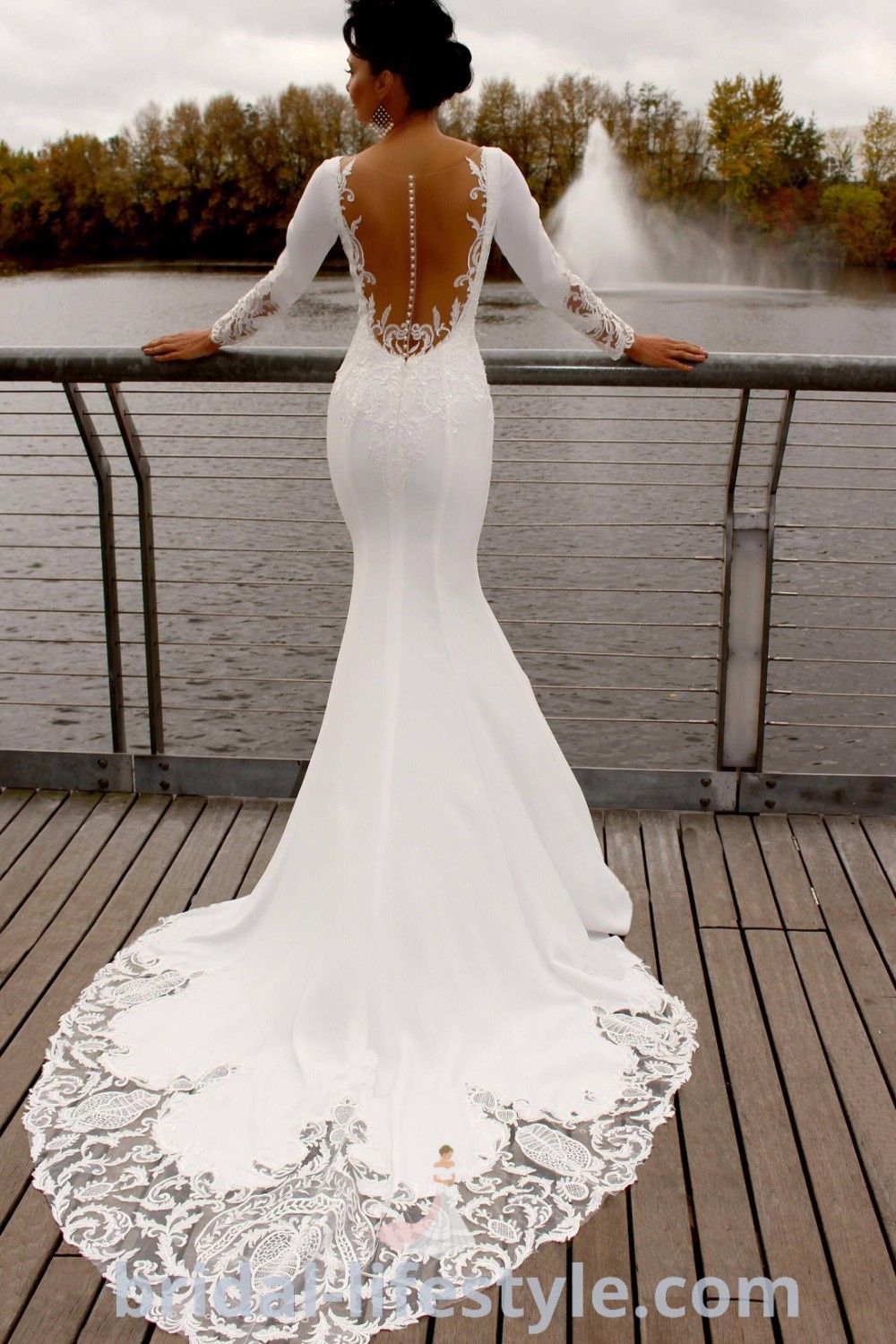 A woman in a white wedding dress with sleeves, embodying elegance and modernity, stands on a pier, looking out at the water. This mermaid dress features exquisite lace details, perfect for the special day. Explore more wedding dress trends and inspiration at bridal-lifestyle.com.