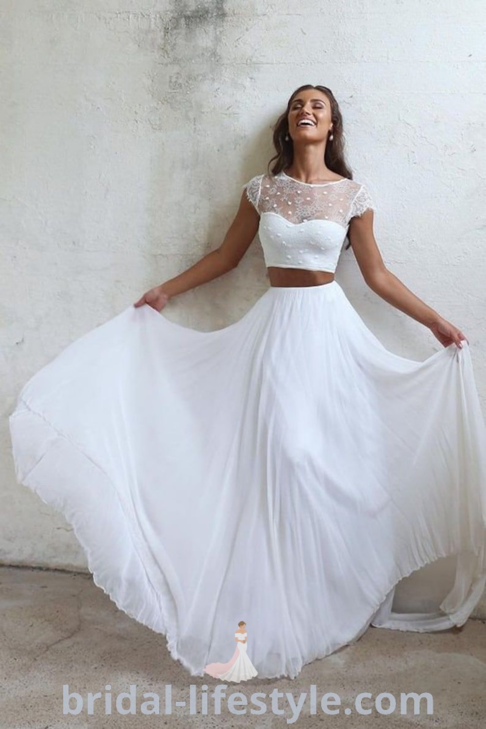 An elegant woman in a white dress poses for the camera, showcasing a rustic barn wedding dress with a lace crop top and two-piece design. This dreamy look blends romantic styles and trends, making it ideal for every bride’s unique taste. Discover the best aesthetic ideas for your special day at bridal-lifestyle.com.