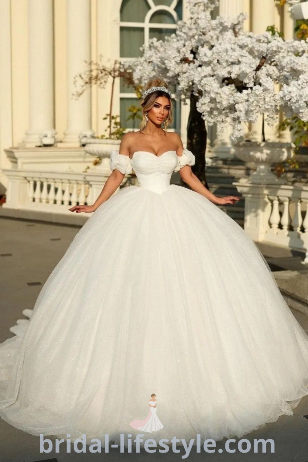 An elegant woman in a white wedding dress with an off-shoulder design, posing confidently with her hands on her hips. This ball gown wedding dress, known for its puffy silhouette, offers a dreamy aesthetic ideal for brides. Discover more stunning styles at bridal-lifestyle.com.