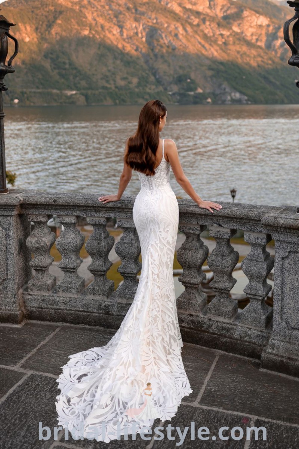 An elegant lace wedding dress with a mermaid silhouette, long train, and breathtaking view of water and mountains, designed for dreamy brides who love refined bridal trends. Discover the best wedding gowns at bridal-lifestyle.com.