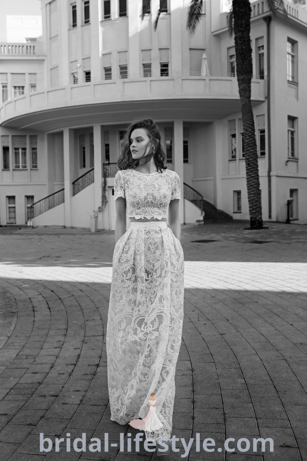 A woman in a long white crop top and skirt wedding outfit stands on a brick walkway near a building, embodying elegance and modern bridal trends. This two piece wedding dress is ideal for brides who want a stylish and unique approach for their special day. Discover more inspiration at bridal-lifestyle.com.