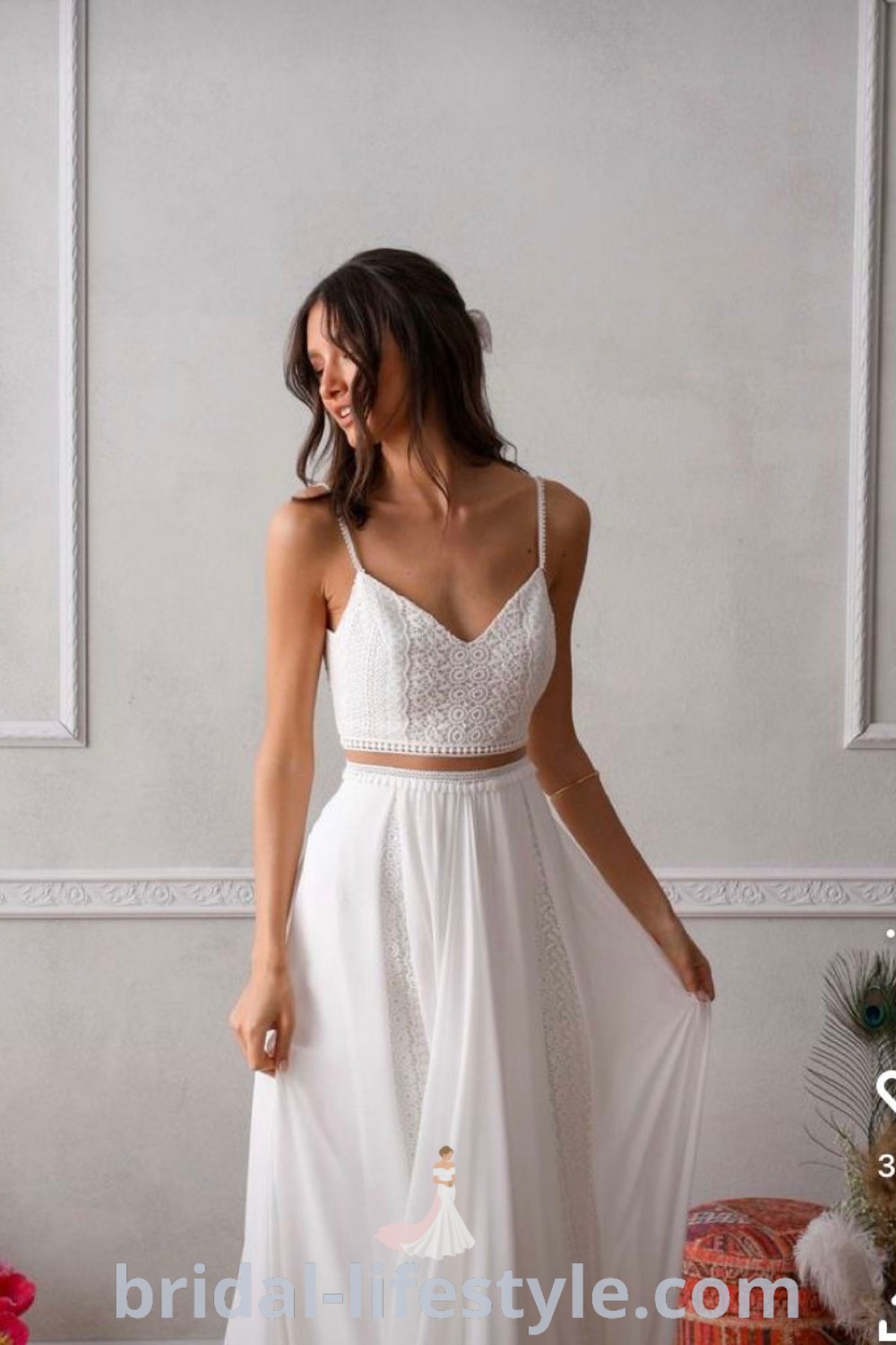 An elegant woman in a white two piece wedding dress standing next to a wall, perfect for boho, beach vow renewals, and tiki weddings. This chic reception dress is ideal for brides seeking dreamy styles that fit every unique taste. Explore more inspiration and trends at bridal-lifestyle.com.