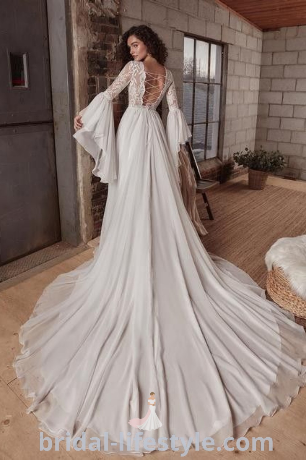 A woman in a long wedding dress with bell sleeves, standing on a wooden floor next to a brick wall, showcasing exquisite Witchy Wedding Dresses from bridal-lifestyle.com. This elegant look combines romantic and Celtic designs for dreamy brides, perfect for unique wedding inspiration on your special day.
