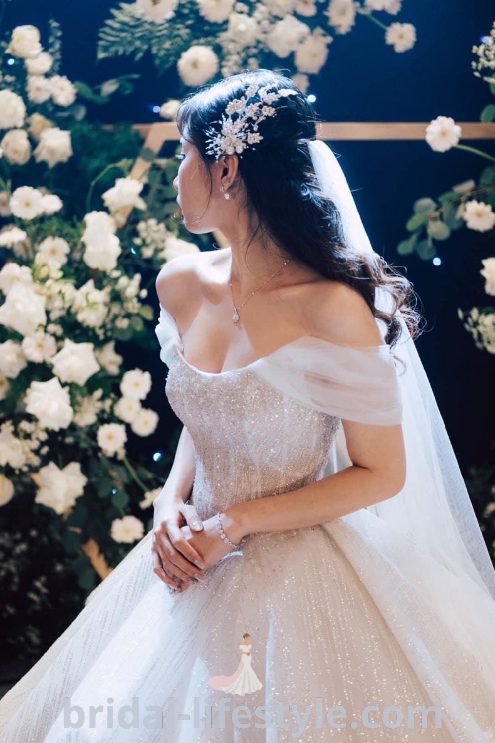 A woman in a fairytale cottage wedding dress with a corset and off-shoulder sleeves, standing next to flowers, embodies a romantic style perfect for June brides. This sparkly ball gown and wedding dress design reflect the best trends in bridal fashion for dreamy brides. Find more inspiration and ideas at bridal-lifestyle.com.