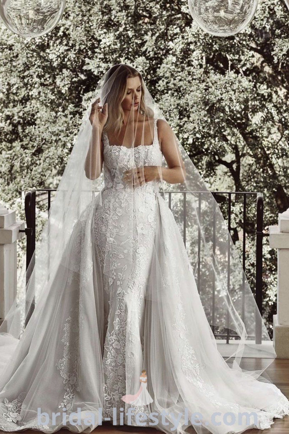 An elegant woman wearing a Galia Lahav ball gown wedding dress and veil stands in front of an archway surrounded by trees, showcasing floral lace details and the latest bridal trends for a romantic atmosphere. Discover more about dreamy wedding dresses at bridal-lifestyle.com.
