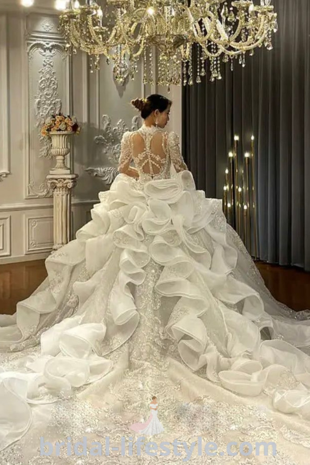 A woman in a beaded ball gown wedding dress stands next to a sparkling chandelier, embodying elegance and romantic vibes for trendy brides. This scene showcases the best inspiration looks for long train wedding dresses and crystal wedding dresses, available at bridal-lifestyle.com.