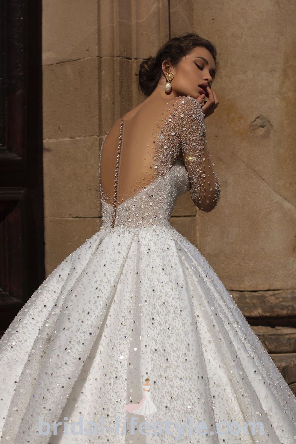 An elegant woman in a sparkly wedding dress with sleeves stands next to a stone wall, showcasing a dreamy bridal look perfect for luxury weddings. This dress embodies trends and aesthetic ideas ideal for every bride