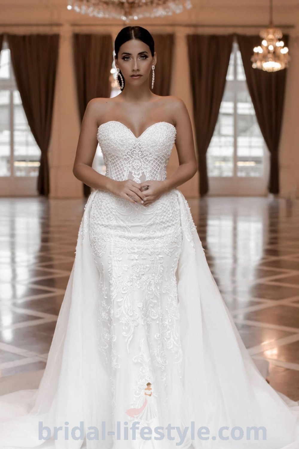 A woman in a stunning white wedding gown stands on a tile floor with a chandelier, embodying elegance and romance. This dress represents the latest wedding gown trends and is perfect for brides seeking inspiration for their special day. Discover more at bridal-lifestyle.com.